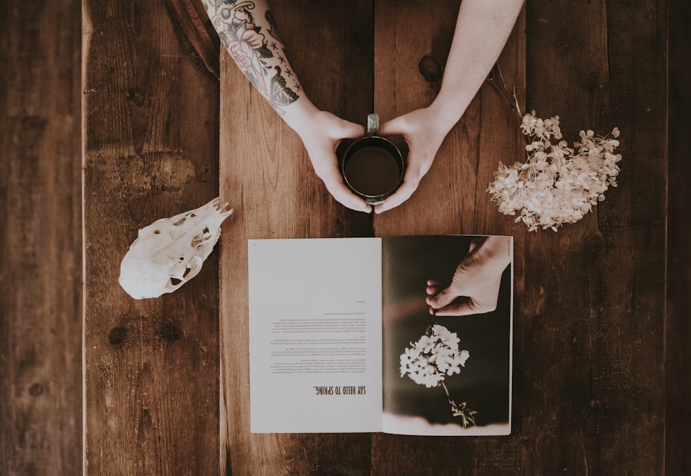 person holding cup with beverage beside white flowers and magazine flat lay photography