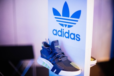 A stylish sneaker is placed beside a panel displaying the Adidas logo, featuring bold blue and white colors. The shoe is predominantly dark with neon green accents and has a modern, sporty design.
