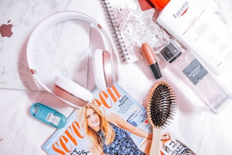 A flat lay photograph features a variety of personal items including over-ear headphones, a magazine, a hairbrush, a lipstick, a bottle of lotion, a fragrance mist, a teal hand sanitizer, and a spiral notebook with a marble-patterned cover lying on a white surface.