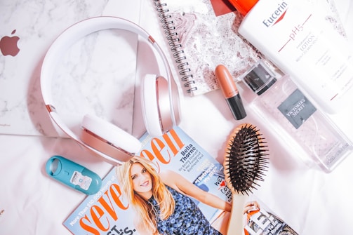 A flat lay photograph features a variety of personal items including over-ear headphones, a magazine, a hairbrush, a lipstick, a bottle of lotion, a fragrance mist, a teal hand sanitizer, and a spiral notebook with a marble-patterned cover lying on a white surface.