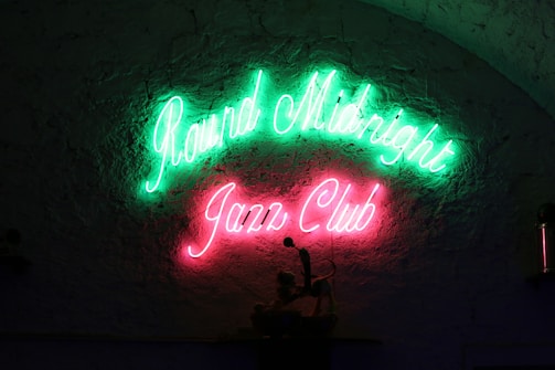 Bright neon signs reading 'Round Midnight Jazz Club' in green and pink are affixed to a textured wall, conveying a lively and inviting atmosphere.