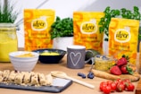 A kitchen countertop features various healthy food items including protein bars on a slate tray, a jar of yellow hummus, a wooden spoon, and a mug with a heart design. Packages of Alver Easy Protein powder are visible in the background. Fresh herbs, a bowl of blueberries, a container with seeds, strawberries, cherry tomatoes, and an avocado are also present, suggesting a focus on natural ingredients.