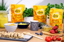 A kitchen countertop features various healthy food items including protein bars on a slate tray, a jar of yellow hummus, a wooden spoon, and a mug with a heart design. Packages of Alver Easy Protein powder are visible in the background. Fresh herbs, a bowl of blueberries, a container with seeds, strawberries, cherry tomatoes, and an avocado are also present, suggesting a focus on natural ingredients.