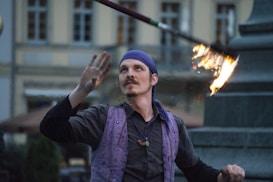 A person skillfully juggling a flaming baton while gazing upward, wearing a purple headscarf and vest against a blurred urban background.