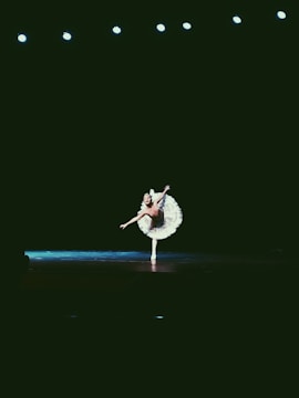 A ballerina performs an elegant pose under stage lights, standing gracefully on one foot. The auditorium is dark, highlighting the dancer's white tutu and the dramatic lighting.