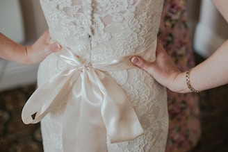 Close-up view of a lace wedding dress with a satin bow tied at the back. Two hands gently rest on the hips, highlighting intricate details of the fabric and the elegance of the outfit. The floral pattern in the background subtly complements the dress.