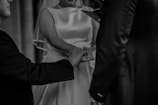 Three people are holding hands, with a focus on the joining of their hands. One person is wearing a light-colored, sleeveless dress, likely a wedding gown, and a veil. The other two are in dark suits. The background is blurred, emphasizing the closeness and significance of this moment.