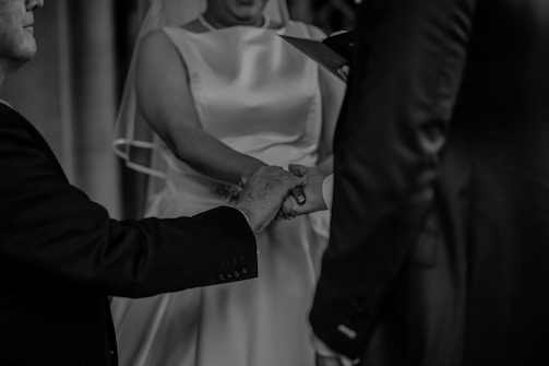 Three people are holding hands, with a focus on the joining of their hands. One person is wearing a light-colored, sleeveless dress, likely a wedding gown, and a veil. The other two are in dark suits. The background is blurred, emphasizing the closeness and significance of this moment.