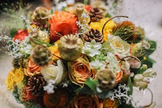 A vibrant arrangement of flowers featuring a variety of blooms including roses in orange, yellow, and white hues. Pinecones and seed pods are interspersed among the flowers, adding texture and a natural touch. Delicate sprigs of greenery and small white flowers are also present, creating a rich and abundant bouquet.
