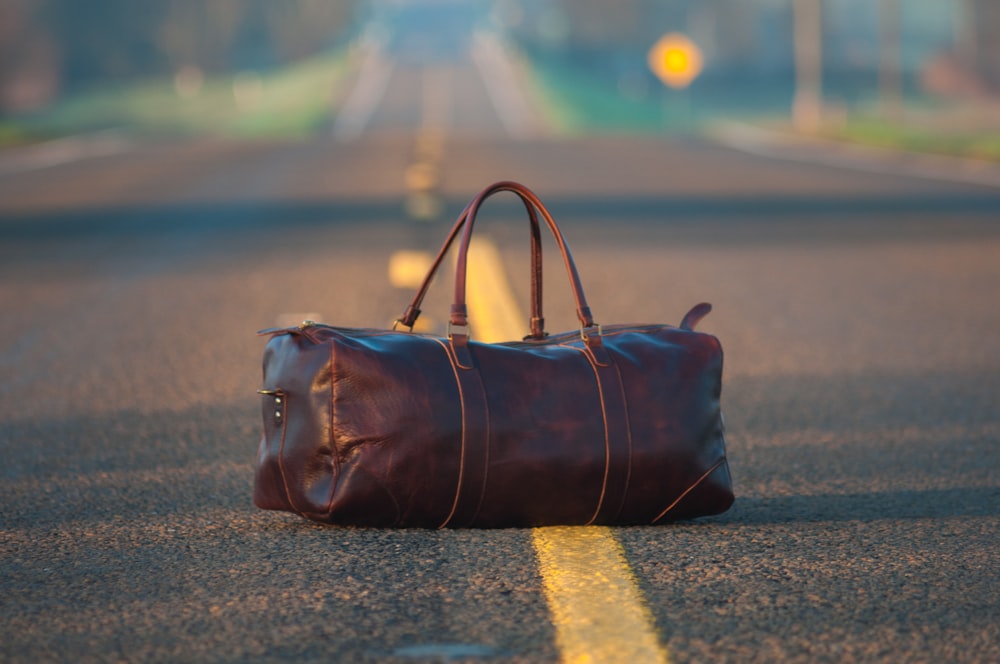 brown leather duffel bag in middle on gray asphalt road