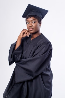 A person wearing a black graduation cap and gown stands confidently against a plain white background, resting their chin on their hand while looking slightly to the side.