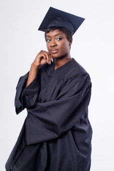 A person wearing a black graduation cap and gown stands confidently against a plain white background, resting their chin on their hand while looking slightly to the side.