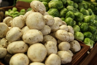 A pile of fresh white mushrooms is in the foreground, while a stack of green Brussels sprouts is visible in the background. The produce is arranged in wooden crates, suggesting a market or grocery setting.