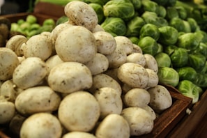 A pile of fresh white mushrooms is in the foreground, while a stack of green Brussels sprouts is visible in the background. The produce is arranged in wooden crates, suggesting a market or grocery setting.