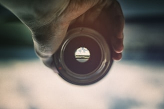 A hand is holding a lens or a circular object, through which a distant landscape with a road and sky is visible, creating a view as if seen through a peephole. The periphery of the lens is blurred, focusing only on the small circular view in the center.