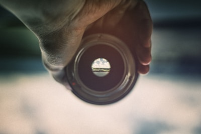 A hand is holding a lens or a circular object, through which a distant landscape with a road and sky is visible, creating a view as if seen through a peephole. The periphery of the lens is blurred, focusing only on the small circular view in the center.