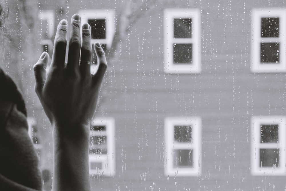 grayscale photo of a pale hand on glass window with rain, other windows visible but out of focus outside. 
