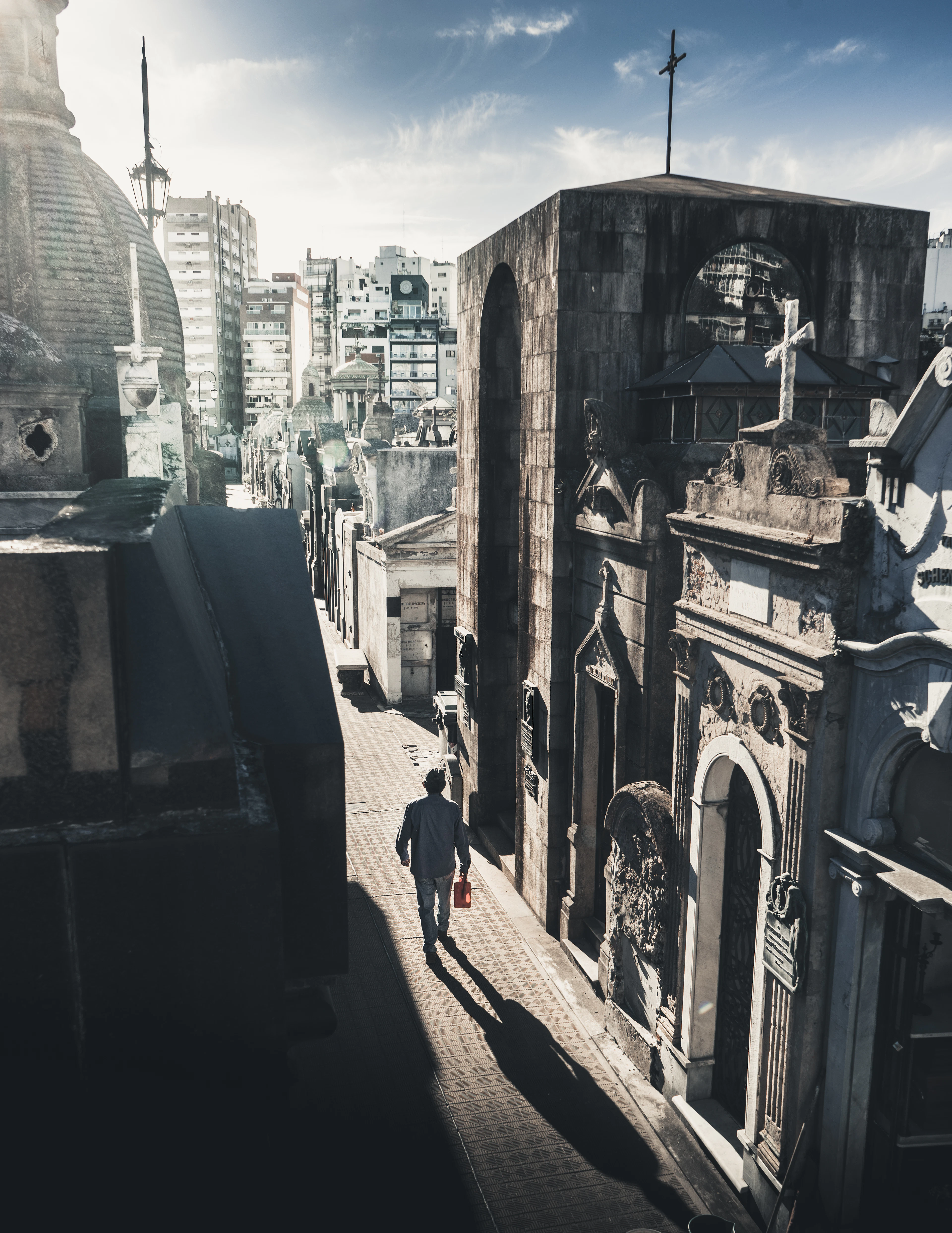 A narrow alley of stone mausoleums and crosses in Recoleta Cemetery, with modern apartment towers rising behind