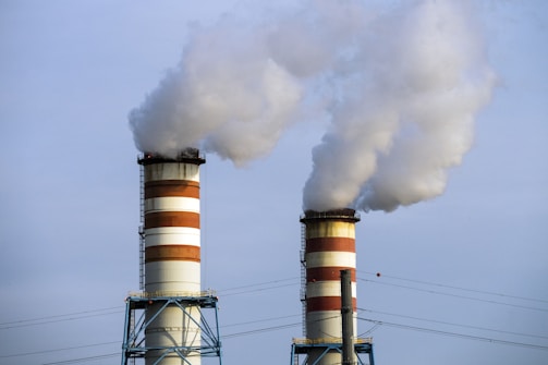 Two industrial chimneys emitting large plumes of smoke into a clear sky. The chimneys are marked with alternating red and white stripes, and they stand on blue metal structures. There are power lines present in the foreground.