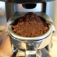 A close-up view of freshly ground coffee in a metal portafilter, ready for brewing in an espresso machine. The coffee grounds appear rich and dark, with a fine texture.