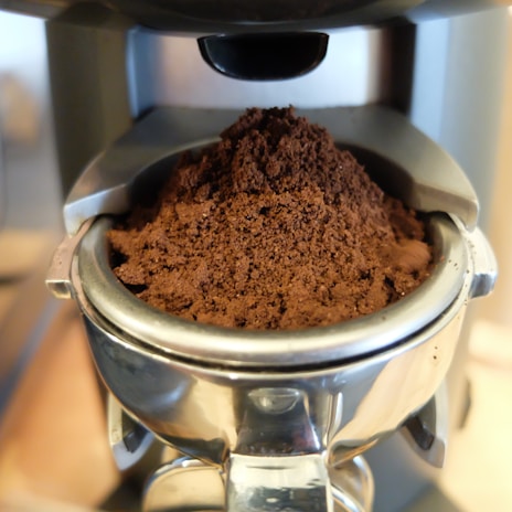 A close-up view of freshly ground coffee in a metal portafilter, ready for brewing in an espresso machine. The coffee grounds appear rich and dark, with a fine texture.
