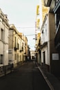 A narrow, empty street lined with closely situated buildings in a European town. The buildings are a mix of older styles with balconies and modern renovations. A sign for a hostel is visible with 'Hostal Rom' written on it. The street appears quiet and slightly shadowed, indicating early morning or late afternoon.
