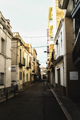 A narrow, empty street lined with closely situated buildings in a European town. The buildings are a mix of older styles with balconies and modern renovations. A sign for a hostel is visible with 'Hostal Rom' written on it. The street appears quiet and slightly shadowed, indicating early morning or late afternoon.