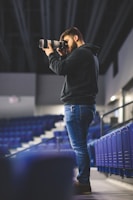 A person stands in an empty stadium, capturing photos with a long lens camera. They are wearing a black hoodie and jeans, focused on their photography. The seats around are unoccupied, creating a serene and quiet atmosphere.