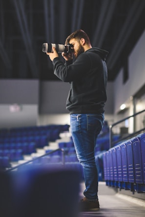 A person stands in an empty stadium, capturing photos with a long lens camera. They are wearing a black hoodie and jeans, focused on their photography. The seats around are unoccupied, creating a serene and quiet atmosphere.