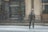 A person dressed in winter clothing is standing on a snow-covered street in front of a building with large columns. Snow is falling heavily, and the street and surrounding areas are blanketed in snow. The person is wearing a jacket, gloves, a hat, and sunglasses.