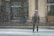 A person dressed in winter clothing is standing on a snow-covered street in front of a building with large columns. Snow is falling heavily, and the street and surrounding areas are blanketed in snow. The person is wearing a jacket, gloves, a hat, and sunglasses.