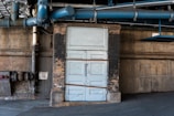 An industrial setting with a large blue pipe running overhead and a weathered door in pastel blue. The surrounding walls are made of aged brick and concrete, with signs of wear and peeling paint. A drain valve sign with red lettering is visible on the left.
