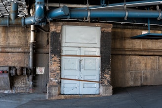 An industrial setting with a large blue pipe running overhead and a weathered door in pastel blue. The surrounding walls are made of aged brick and concrete, with signs of wear and peeling paint. A drain valve sign with red lettering is visible on the left.