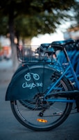 A blue bicycle with a basket on the rear is parked on a street. The bike is part of a rental service called ClujBike, indicated by the logo on its back wheel cover. Nearby, other bicycles can be seen, suggesting a bike-sharing station. The background features blurry trees and a sidewalk, with a warm, soft light.