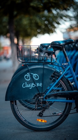 A blue bicycle with a basket on the rear is parked on a street. The bike is part of a rental service called ClujBike, indicated by the logo on its back wheel cover. Nearby, other bicycles can be seen, suggesting a bike-sharing station. The background features blurry trees and a sidewalk, with a warm, soft light.