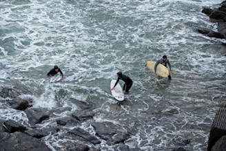 Three surfers are in the water close to a rocky shore, wearing wetsuits and holding surfboards. The waves are choppy, and the scene captures them either preparing to enter or exit the water.