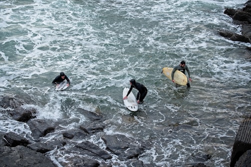 Three surfers are in the water close to a rocky shore, wearing wetsuits and holding surfboards. The waves are choppy, and the scene captures them either preparing to enter or exit the water.