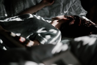 The image depicts a person lying in bed with soft light partially illuminating their face and upper body. The shadows create a dappled effect across the sheets and the person's skin. The atmosphere appears calm and serene.