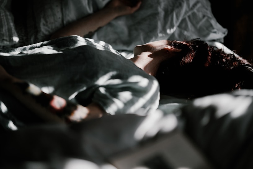 The image depicts a person lying in bed with soft light partially illuminating their face and upper body. The shadows create a dappled effect across the sheets and the person's skin. The atmosphere appears calm and serene.