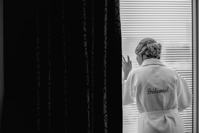 A woman wearing a robe with 'Bridesmaid' embroidered on the back stands looking out a window. Her hair is styled elegantly, and she appears to be gazing thoughtfully outside. The setting is serene and private with dark curtains on the left side offering a contrast to the bright window light.