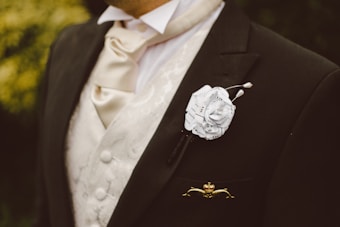 A close-up of a person wearing formal attire. The individual is dressed in a black suit jacket with a white patterned vest underneath. A cream cravat is elegantly tied around the neck. There is a distinctive white boutonniere made from sheet music attached to the lapel.