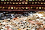 A large collection of assorted coins scattered across a surface, featuring different sizes and metallic shades. The coins have a weathered appearance, some reflecting light, and others appearing dull. In the background, there is an out-of-focus row of round objects resembling the coins, enhancing the depth of the image.