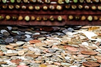 A large collection of assorted coins scattered across a surface, featuring different sizes and metallic shades. The coins have a weathered appearance, some reflecting light, and others appearing dull. In the background, there is an out-of-focus row of round objects resembling the coins, enhancing the depth of the image.