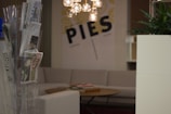 A cozy indoor setting featuring a rack with various newspapers and magazines, a comfortable beige couch, and a round wooden coffee table with books or magazines. A modern chandelier with spherical bulbs hangs above, and the wall displays a large graphic with the word 'PIES'. There's a green plant in a white planter on the right side.