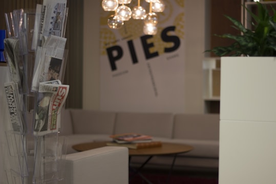 A cozy indoor setting featuring a rack with various newspapers and magazines, a comfortable beige couch, and a round wooden coffee table with books or magazines. A modern chandelier with spherical bulbs hangs above, and the wall displays a large graphic with the word 'PIES'. There's a green plant in a white planter on the right side.