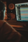 A dimly lit sound studio with an audio mixing console, a computer screen displaying audio tracks, and a speaker in the background. The scene has a warm, focused ambiance with a person partially visible, likely working on audio production.