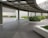 A modern outdoor area with a covered walkway featuring a transparent roof. The space is bordered by neatly trimmed green hedges and surrounded by gray concrete structures and paved tiles. Buildings with a minimalist design are visible in the background.