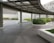 A modern outdoor area with a covered walkway featuring a transparent roof. The space is bordered by neatly trimmed green hedges and surrounded by gray concrete structures and paved tiles. Buildings with a minimalist design are visible in the background.
