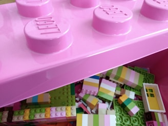A close-up view of a pink storage box designed to look like a large LEGO brick. The box is open, revealing an assortment of multicolored LEGO blocks inside, primarily composed of pastel colors such as light green, pink, yellow, and white. Some blocks are partially constructed or connected.