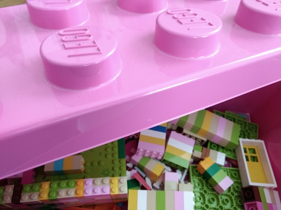 A close-up view of a pink storage box designed to look like a large LEGO brick. The box is open, revealing an assortment of multicolored LEGO blocks inside, primarily composed of pastel colors such as light green, pink, yellow, and white. Some blocks are partially constructed or connected.